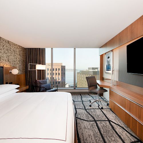A modern hotel room features a large bed, a flat-screen TV, a desk and chair by the window, and a view of the city and water.