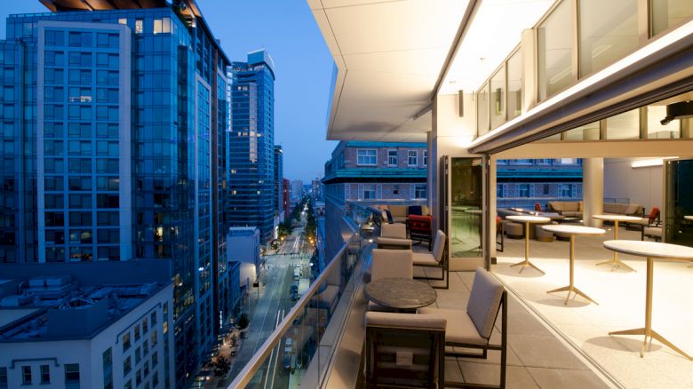 The image shows a modern, high-rise balcony overlooking a cityscape at dusk, with outdoor seating and a glass railing enhancing the view.