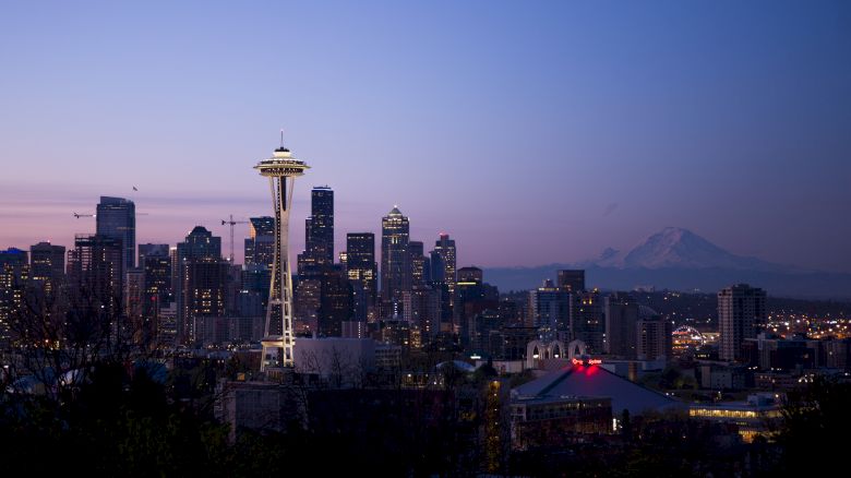 The image shows a twilight view of the Seattle skyline with the Space Needle and Mount Rainier in the background. The city lights are starting to illuminate.