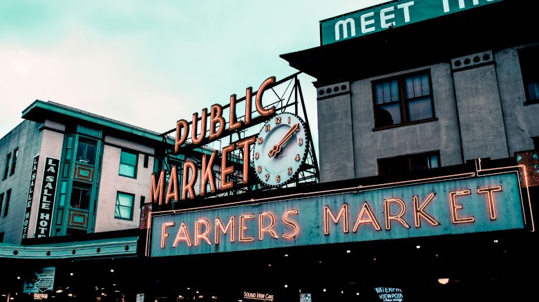 This image shows the iconic neon signs of the Public Market and Farmers Market at Pike Place Market in Seattle, with buildings in the background.