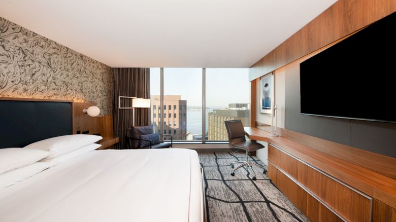 A modern hotel room with a large bed, flat-screen TV, desk, chair, and a view of buildings through a large window, providing a clean, minimalist look.