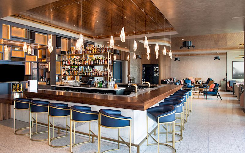 The image shows a modern bar with a wooden counter, blue bar stools, hanging lights, and a well-stocked liquor shelf in a spacious, stylish setting.