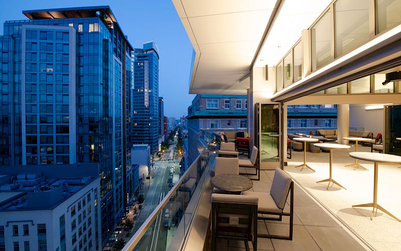 A modern balcony with seating overlooks a city street with tall buildings, illuminated in the evening.