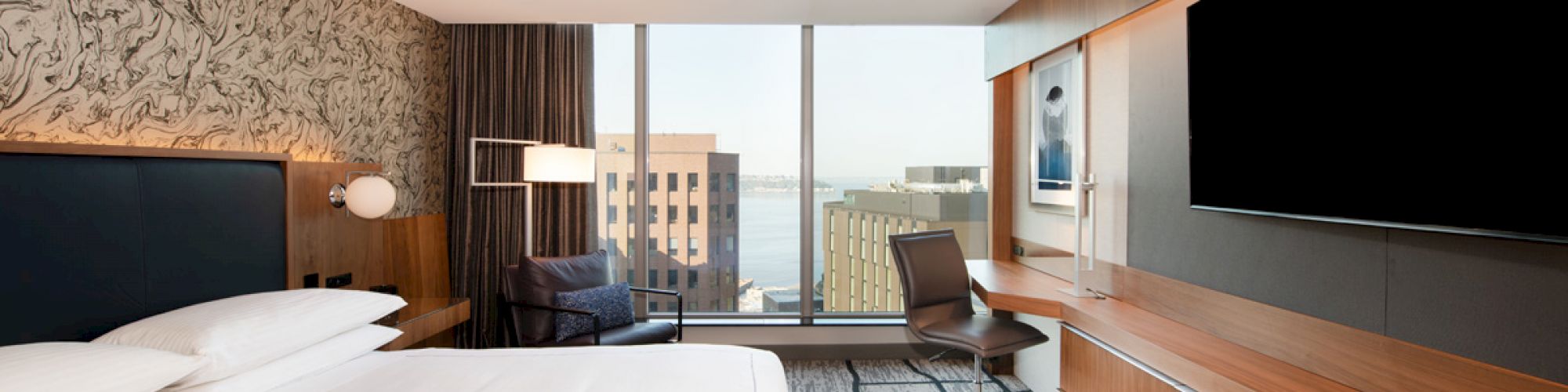 A modern hotel room with a king-sized bed, large wall-mounted TV, desk, chair, and window with a cityscape view in the background.