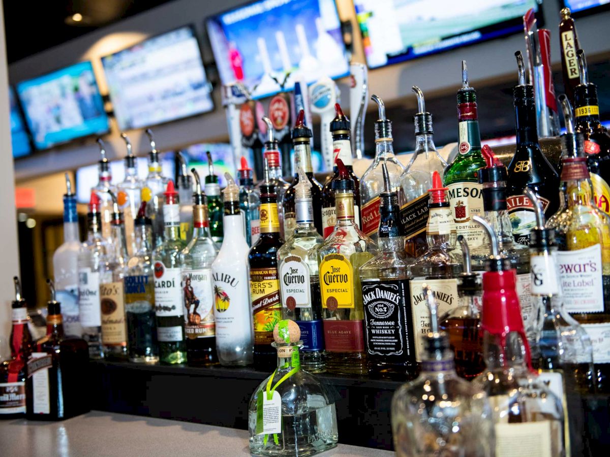 A bar counter filled with various liquor bottles, including tequila, vodka, whiskey, rum, and more, with multiple TV screens in the background.