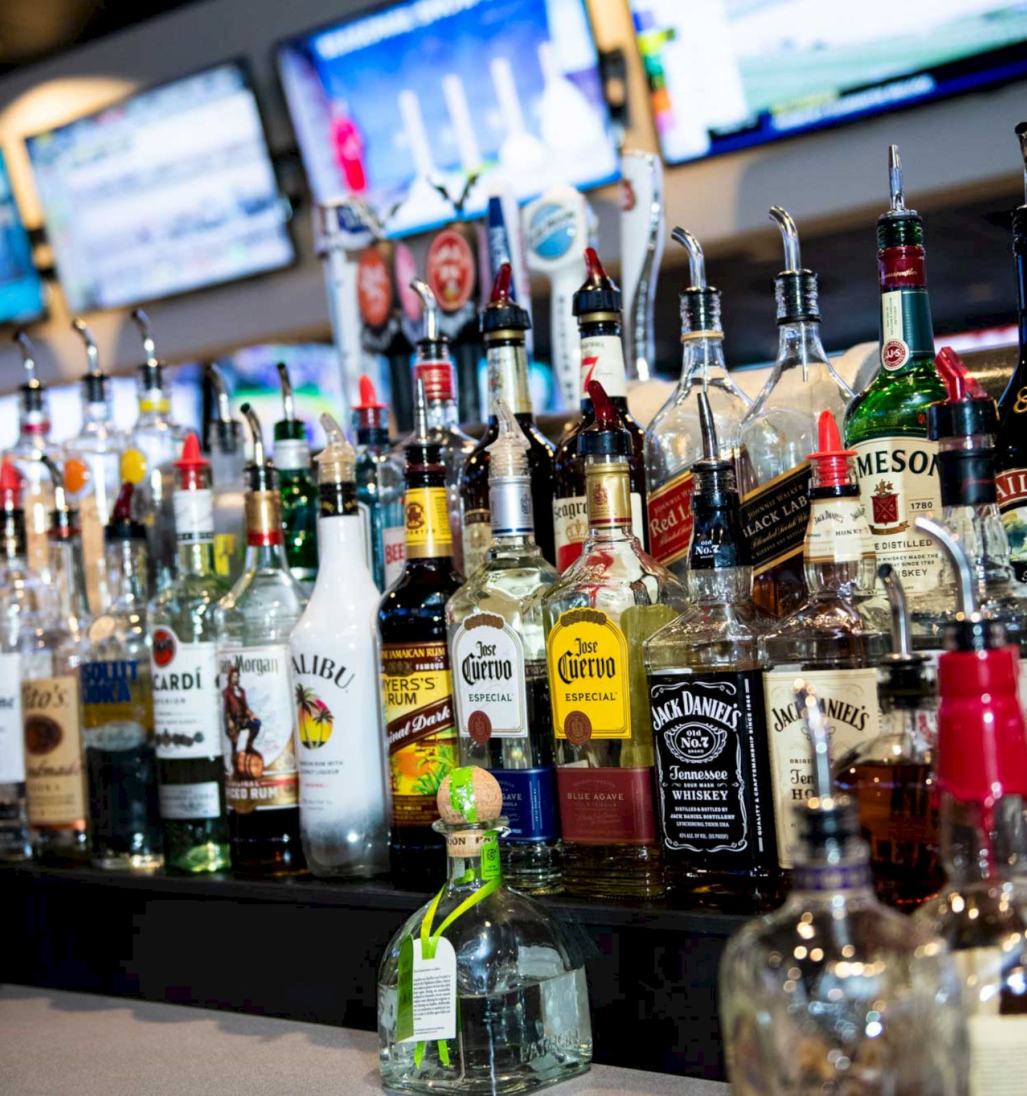 A bar counter filled with various liquor bottles, including tequila, vodka, whiskey, rum, and more, with multiple TV screens in the background.