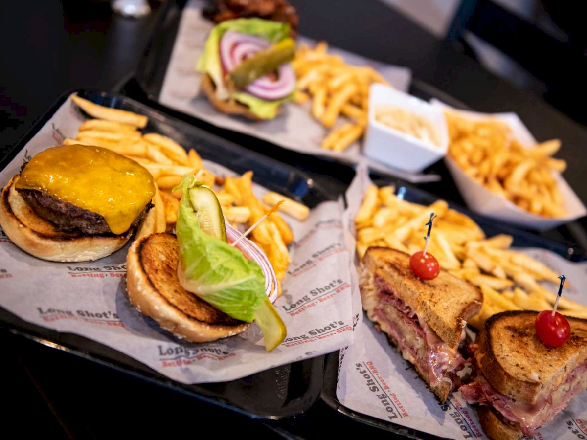 The image shows trays with cheeseburgers, sandwiches, and servings of fries, garnished with pickles and vegetables on a dark table ending the sentence.