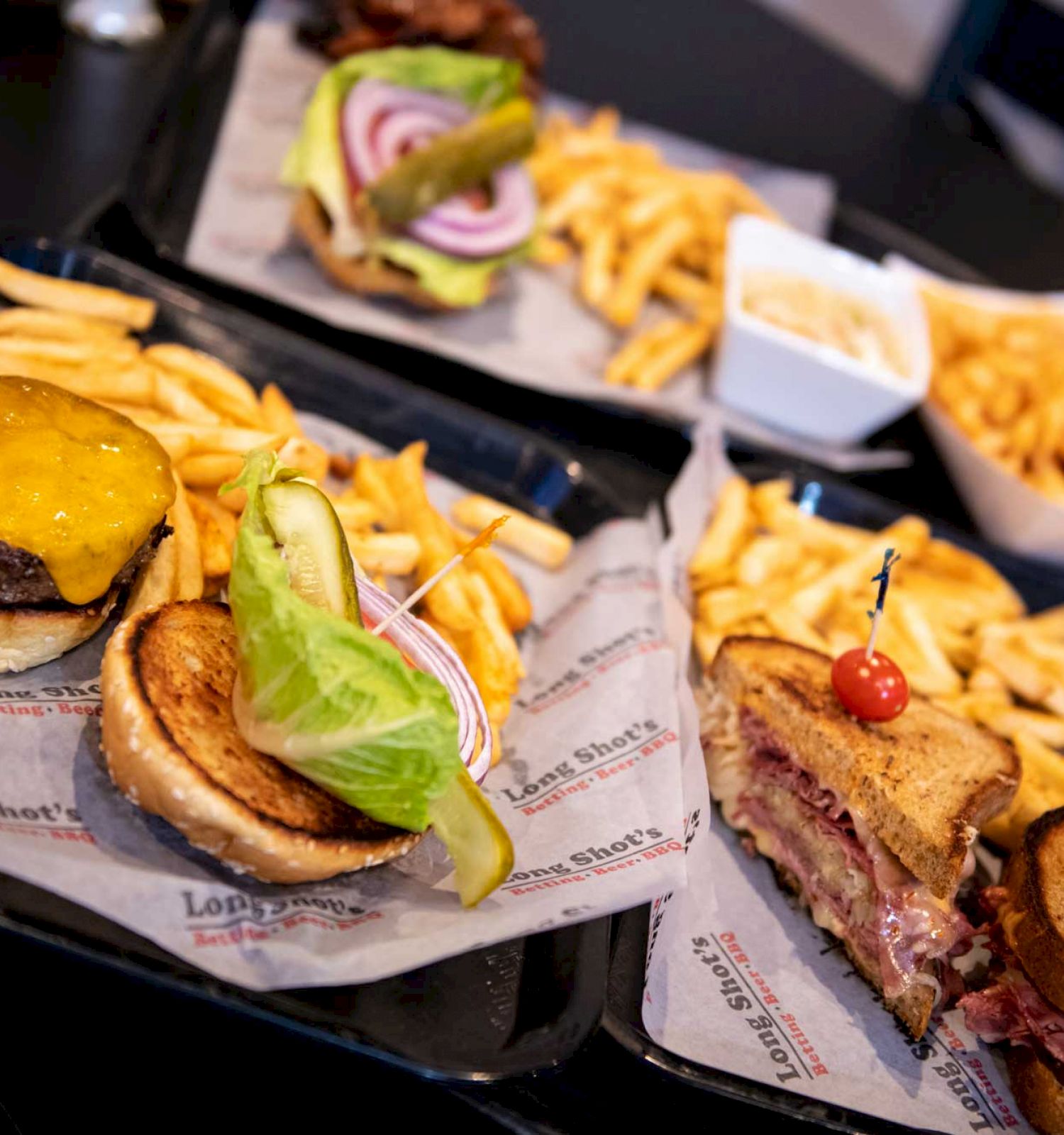 The image shows trays with cheeseburgers, sandwiches, and servings of fries, garnished with pickles and vegetables on a dark table ending the sentence.