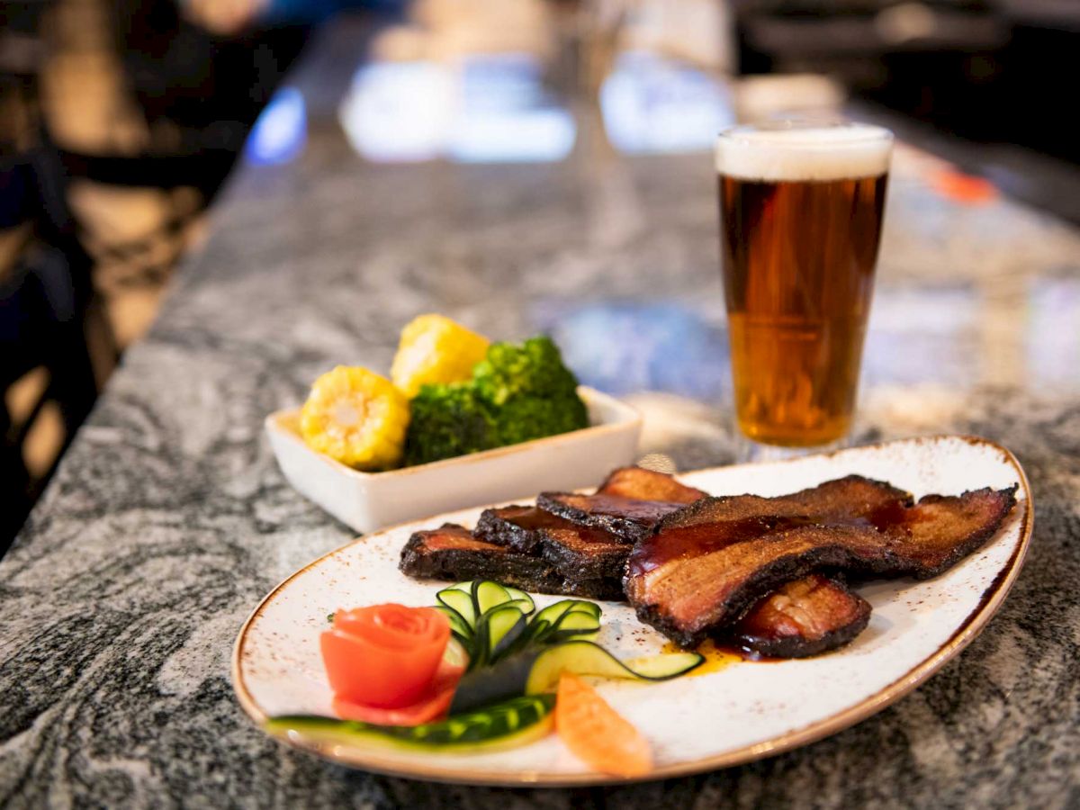 The image shows a plate of roasted meat with vegetables and a glass of beer on a marble countertop, with a side dish of broccoli and corn.