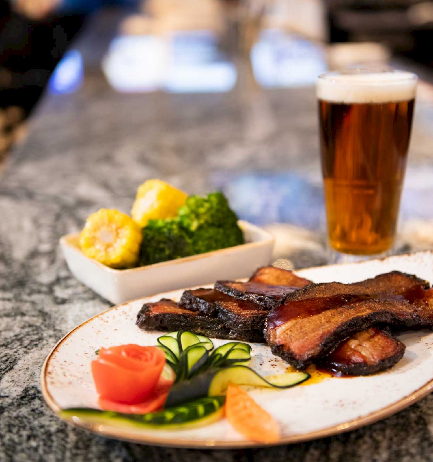 The image shows a plate of roasted meat with vegetables and a glass of beer on a marble countertop, with a side dish of broccoli and corn.