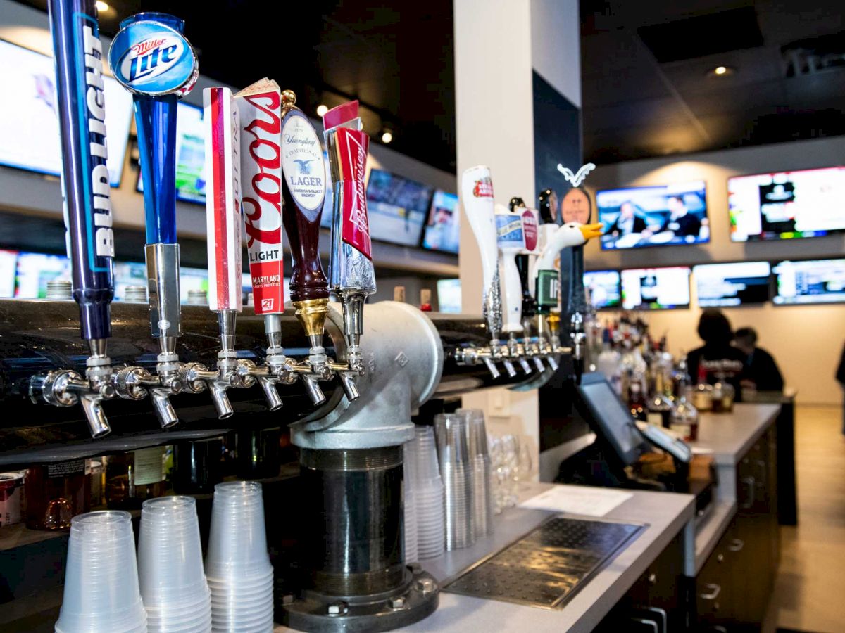 This image shows a bar with several beer taps, plastic cups, and multiple TVs in the background. The setting appears to be a sports bar.