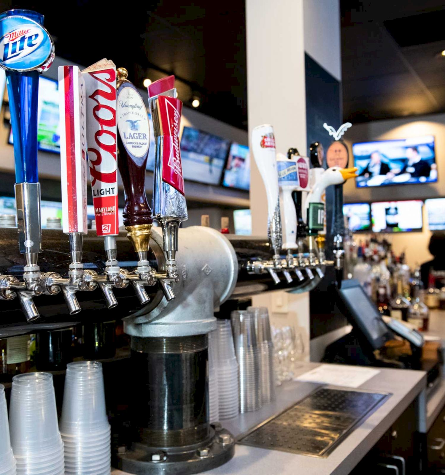 This image shows a bar with several beer taps, plastic cups, and multiple TVs in the background. The setting appears to be a sports bar.