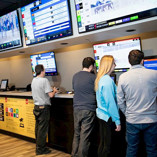 People stand at a counter with betting terminals under multiple screens displaying sports information and odds, likely in a sportsbook.