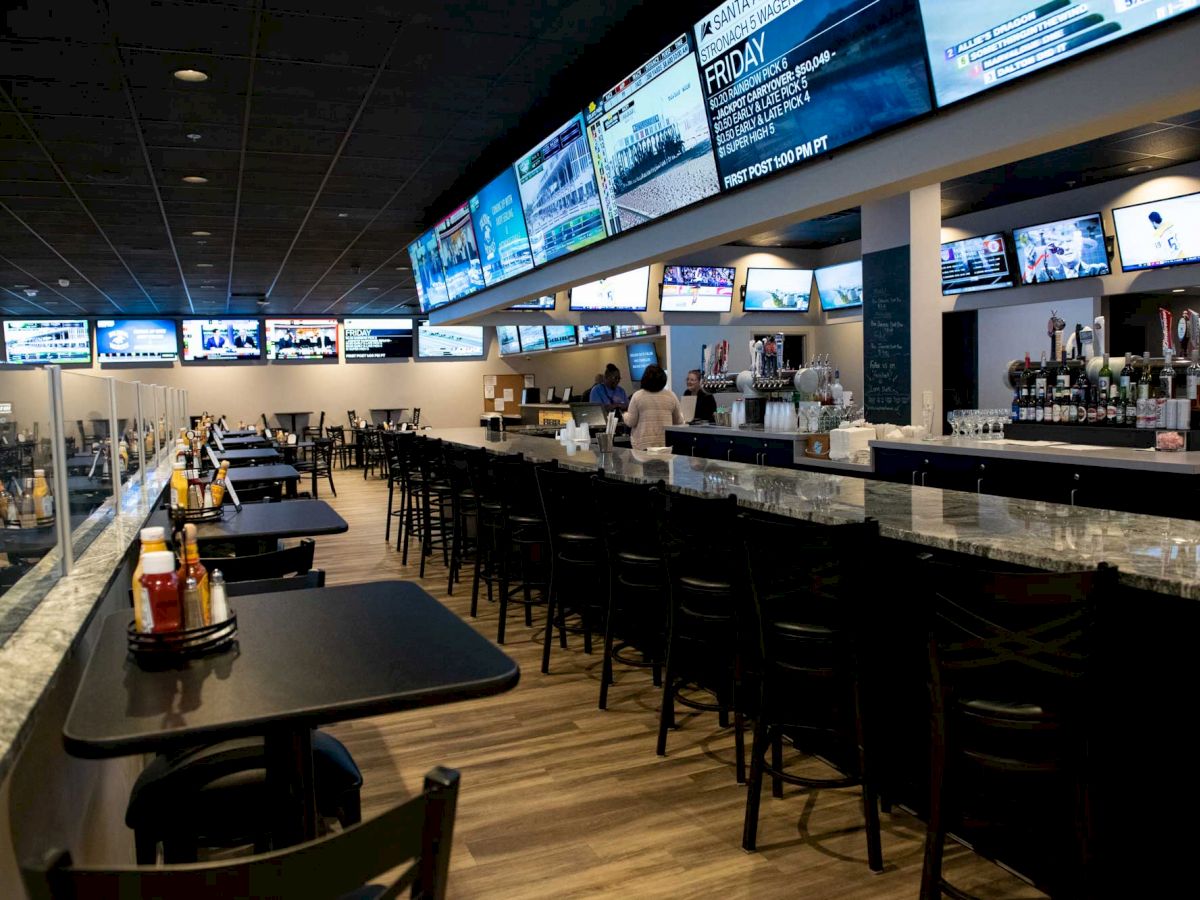 The image shows an empty sports bar with multiple TVs, a long bar counter with stools, and tables set with condiments.